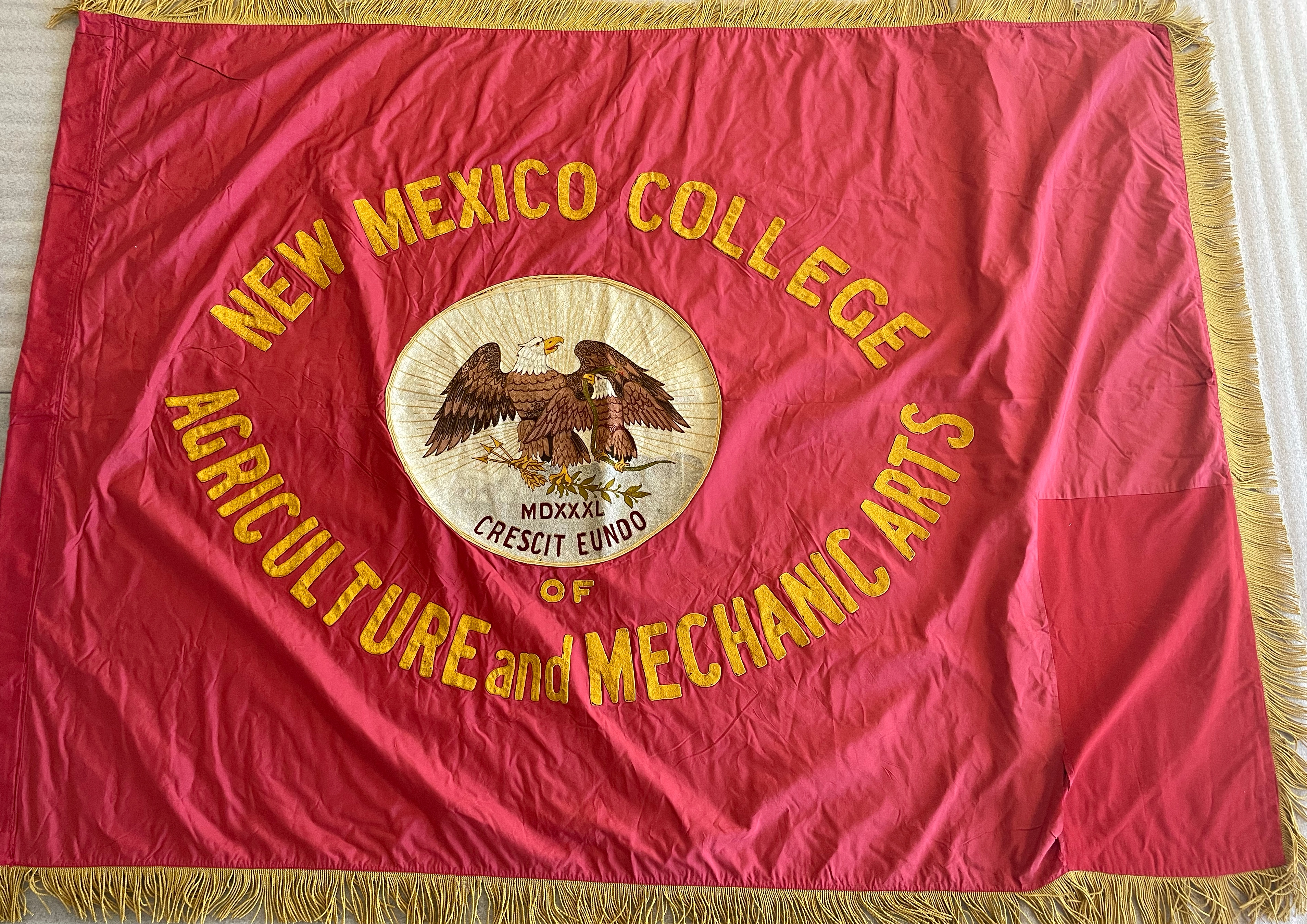 Red New Mexico College of Agriculture and Mechanic Arts flag/banner with seal of state of New Mexico in center and gold lettering