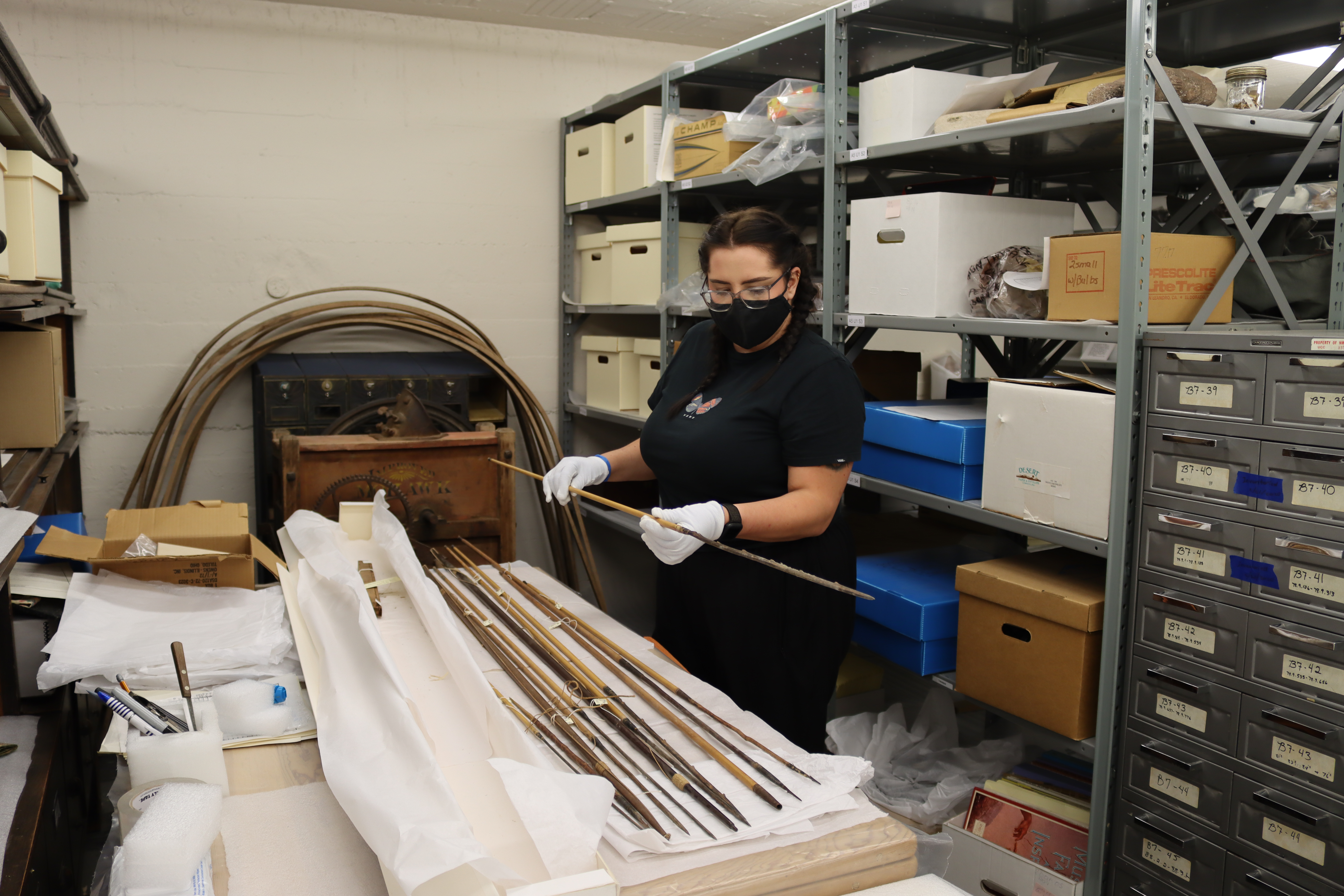 Carly Johston holds a thin spear while inventorying it for project. She is in the basement collections storage, wearing a black shirt and black mask. 